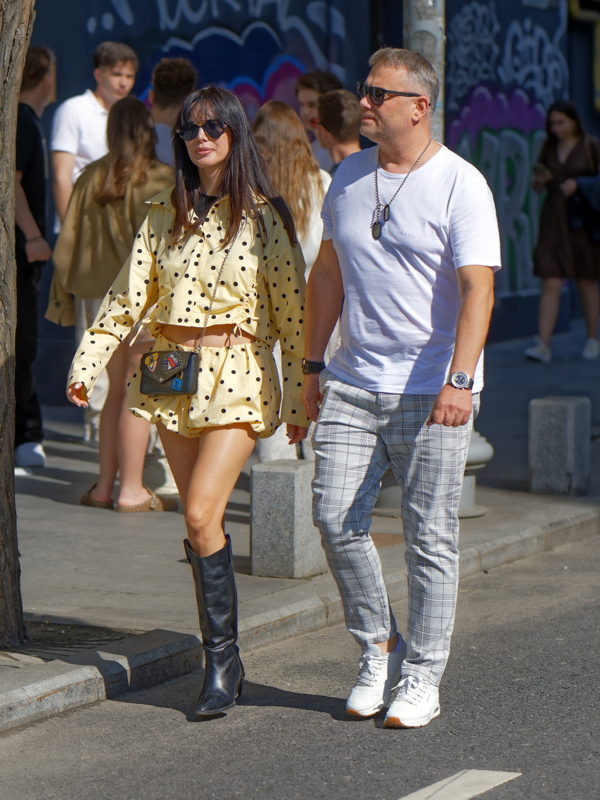 Woman wearing a yellow polka dot outfit with a matching bag and black knee-high boots, walking alongside a man in a white t-shirt and plaid trousers